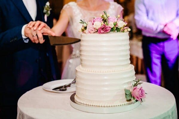 Brautpaar schneidet eine mehrstöckige weisse Hochzeitstorte mit Blumendekoration an – typisches Bild einer Hochzeitstorte in der Schweiz.