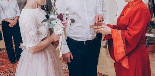 Bei einer Trauung greift ein Paar nach einem Ring, während eine Frau in rotem Gewand eine kleine Schale hält; Gäste im Hintergrund.