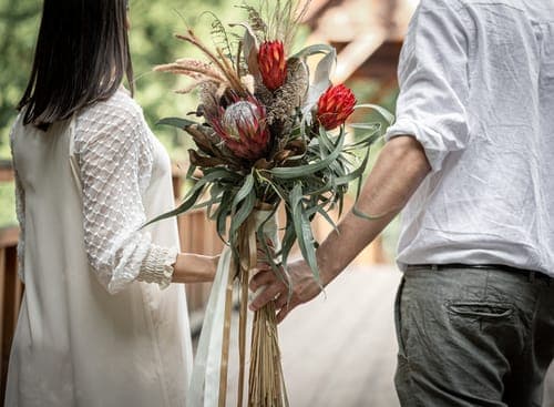 Zwei Personen reichen sich auf einem Holzsteg einen grossen, exotischen Blumenstrauss mit roten Blüten und langen Blättern.