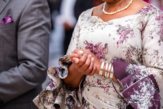 Eine festlich gekleidete Frau in cremefarbenem Kleid mit violettem Muster, Perlenkette und Armreifen; ihre Hände sind gefaltet.