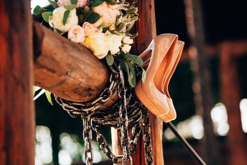 Zwei beigefarbene High Heels hängen an einer Holzbalken-Konstruktion neben einem zarten Blumenarrangement und Ketten.