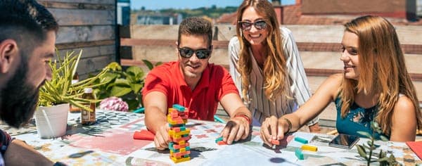 Freundesgruppe spielt Jenga auf der Terrasse; ein Mann zieht einen Holzklotz aus dem Turm, alle schauen gespannt zu.
