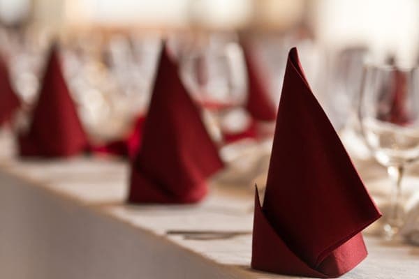 Dunkelrote, spitz gefaltete Servietten in Reihe auf festlich gedeckter Tafel mit Weingläsern im Hintergrund.