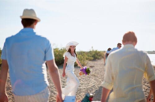 Braut in weissem Kleid bei ihrer Strandhochzeit, von Gästen umgeben, geht über den Sand zum Altar.