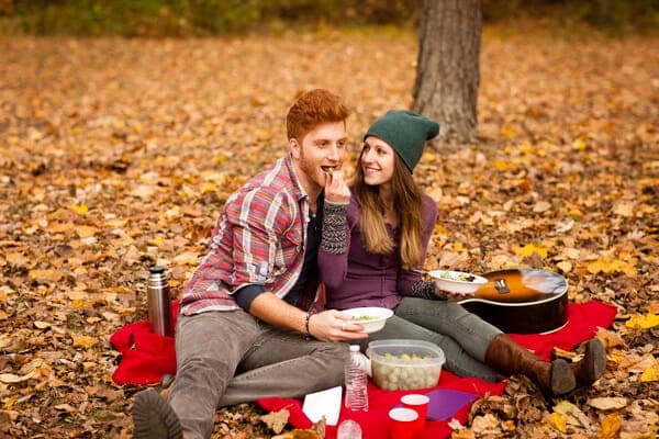 Paar macht Picknick auf einer roten Decke im Herbstlaub; sie füttert ihn, daneben liegen eine Gitarre und viele Herbst-Blätter.