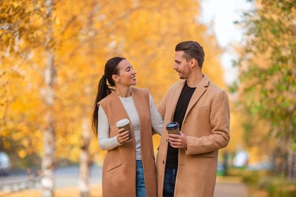 Paar mit braunem Herbst Outfit spaziert durch einen Herbstpark, spricht miteinander und hält Kaffee zum Mitnehmen; goldene Bäume im Hintergrund.