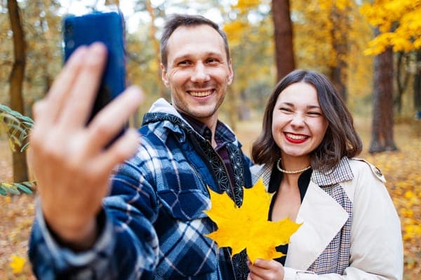 Lächelndes Paar macht ein Selfie im Herbstwald; die Frau hält ein grosses gelbes Ahornblatt in die Kamera.