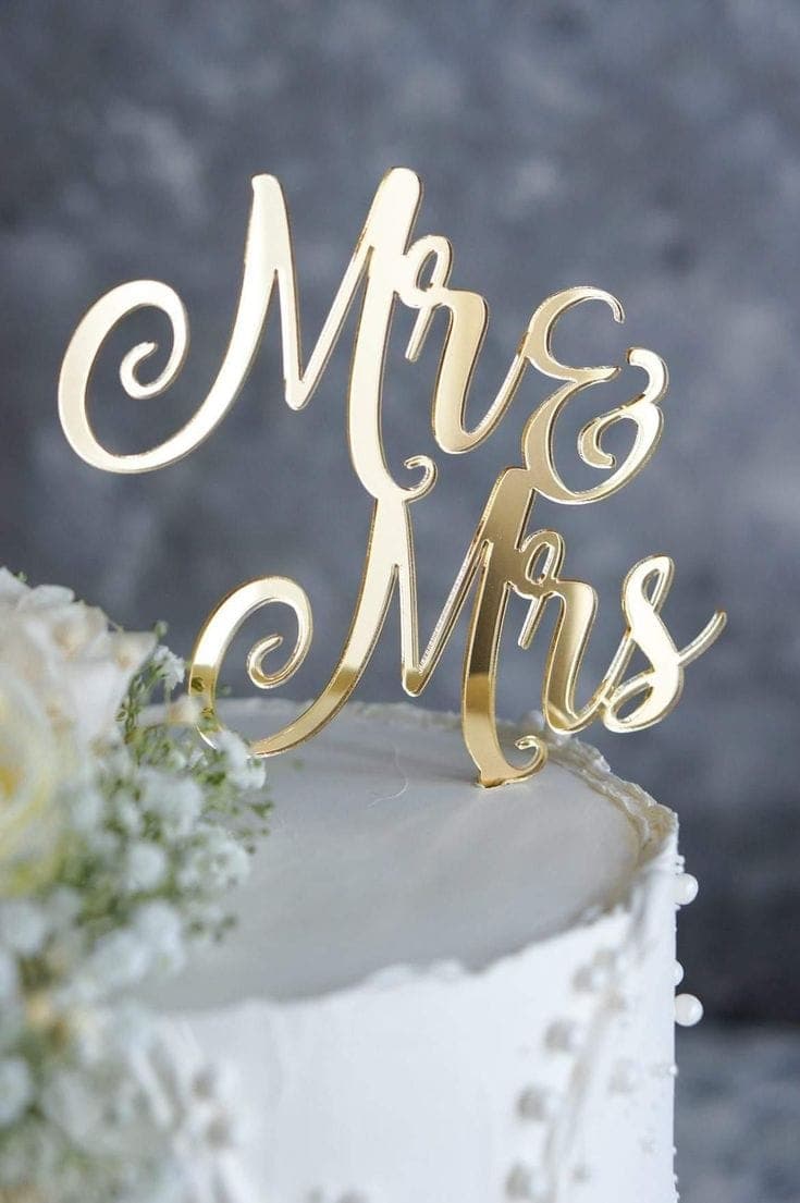Ein glänzender, goldener "Mr & Mrs" Cake Topper aus Acryl, der eine festliche Hochzeit Cake mit zarten weissen Blumen schmückt.