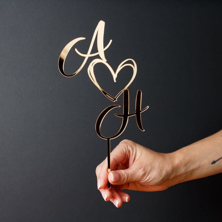 Eine Hand hält einen filigranen Cake Topper Hochzeit personalisiert aus goldenem Acryl, der zwei Initialen mit einem Herz verbindet.