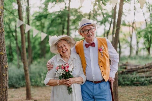 Fröhliches älteres Paar im Wald bei einer Feier: sie im weissen Kleid und Hut mit Blumenstrauss, er mit Strohhut, roter Fliege und orangefarbener Weste.