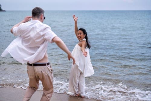 Glückliches Brautpaar bei einer Strandhochzeit, Händchen haltend am Wasserrand.