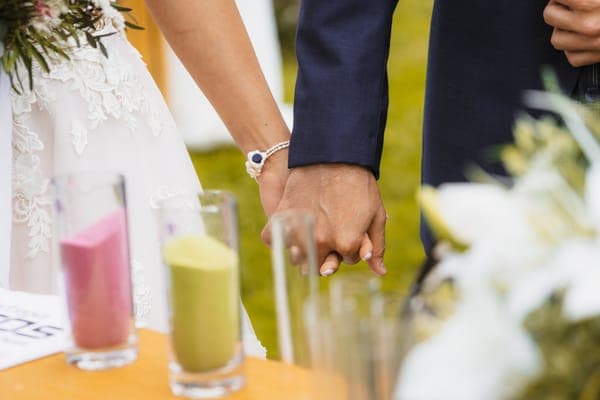 A bride and groom holding hands during an outdoor sand ceremony, with rings, glasses and flowers in the foreground