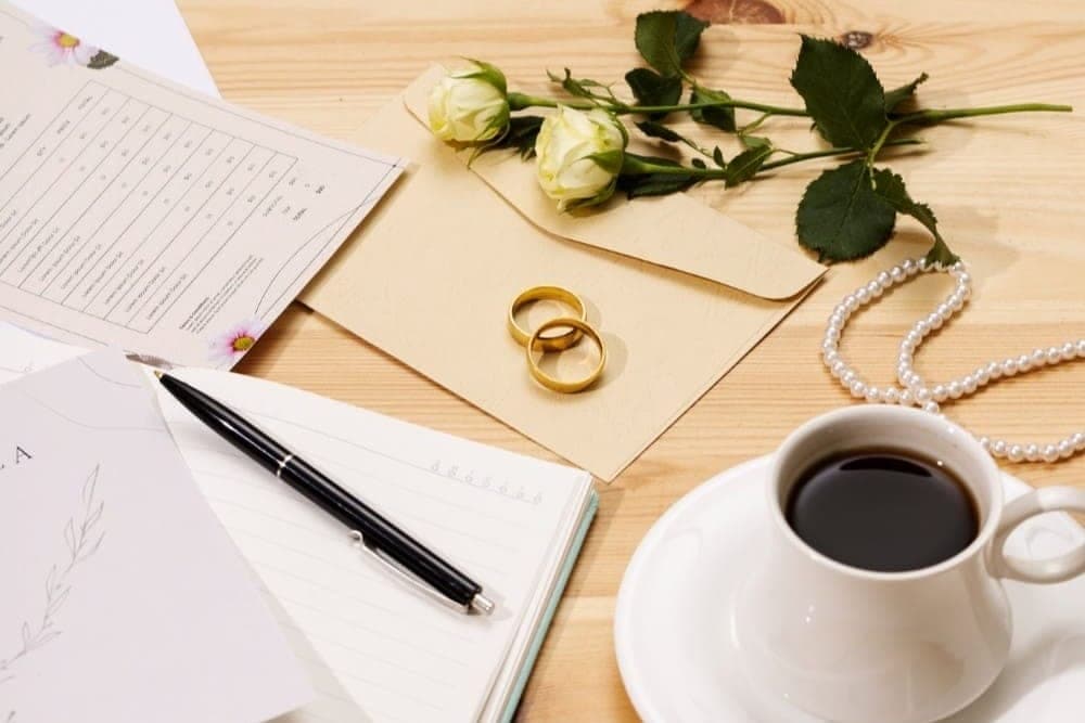 Tisch mit Listen, Stift, zwei Eheringen auf einem Briefumschlag, weissen Rosen und einer Tasse Kaffee