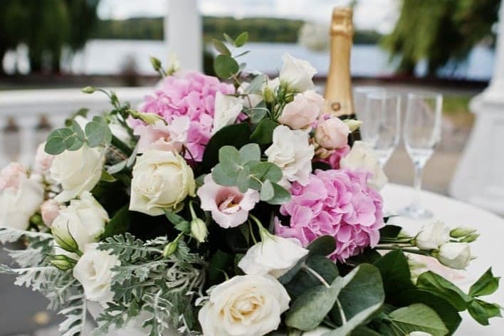 Blumengesteck mit weissen Rosen und rosa Hortensien auf einem Tisch, dahinter eine Flasche Champagner und zwei Gläser