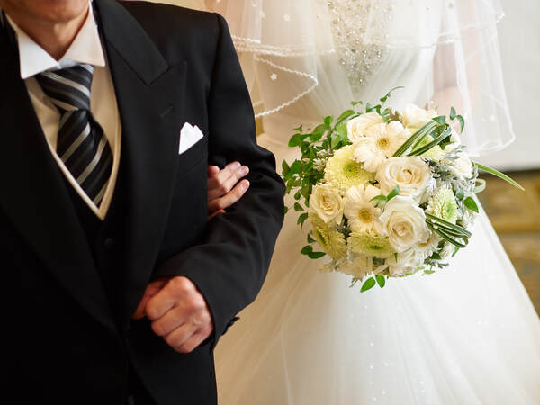 Braut in weissem Spitzenkleid hält einen weissen Blumenstrauss und hakt sich beim Begleiter im schwarzen Anzug unter.