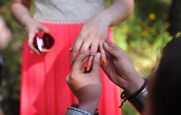 Detailaufnahme eines kreativen Heiratsantrags im Freien: Ein Mann in Rüstung steckt einer Frau in einem korallfarbenen Kleid einen Verlobungsring mit lila Stein an den Finger.