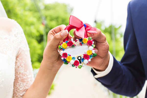 Persönliche Glückwünsche zur Hochzeit: So findest du die perfekten Worte Hochzeitswünsche, Hochzeitsblog Hände eines Brautpaares halten gemeinsam ein herzförmiges, mit bunten Blumen verziertes Liebesschloss mit rosa Schleife.