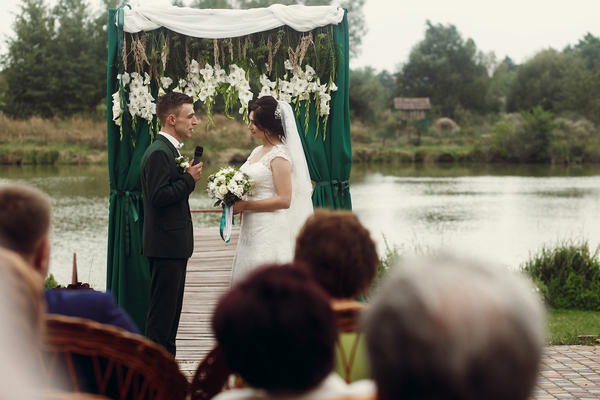 Eiserne Hochzeit: Die beeindruckende Feier der Stärke nach 65 gemeinsamen Jahren Hochzeitsblog Brautpaar bei freier Trauung am See unter grünem Traubogen mit weissen Blumen, Gäste sitzen im Vordergrund.