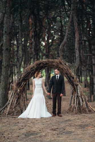 Hölzerne Hochzeit: Der grosse Guide zum Jubiläum aus Holz Hochzeitsjubiläen, Hochzeitsblog Brautpaar steht Händchen haltend im Wald unter einem grossen Bogen aus Ästen; sie trägt ein weisses Kleid, er Anzug.
