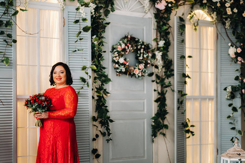 Rubinhochzeit: Der ultimative Guide für einen unvergesslichen 40. Hochzeitstag Hochzeitsjubiläen, Hochzeitsblog Frau im roten Spitzenkleid hält einen Blumenstrauss und steht vor einer dekorierten Tür mit Blumenkranz und Efeu.