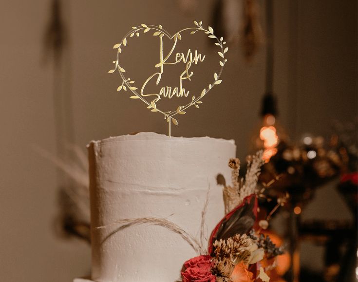 Ein eleganter, goldener Cake Topper Hochzeit personalisiert mit den Namen "Kevin & Sarah" in einem Herz aus Blättern, der eine schlichte, weisse Hochzeit Cake krönt.